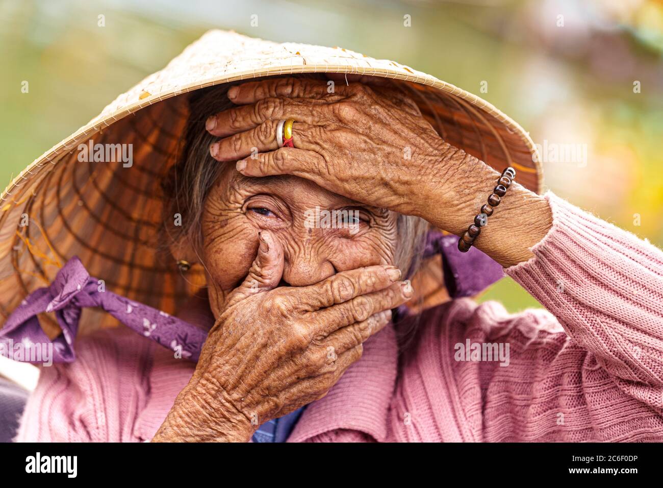 Old Vietnamese woman in violet sweater puts her hands to her face to cover her mouth and forehead. Stock Photo