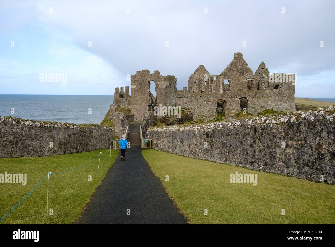 Lady in light blue coat walks down the entrance path towards the spectacular ruins of Dunluce Castle near Portrush, Co. Antrim, Northern Ireland Stock Photo