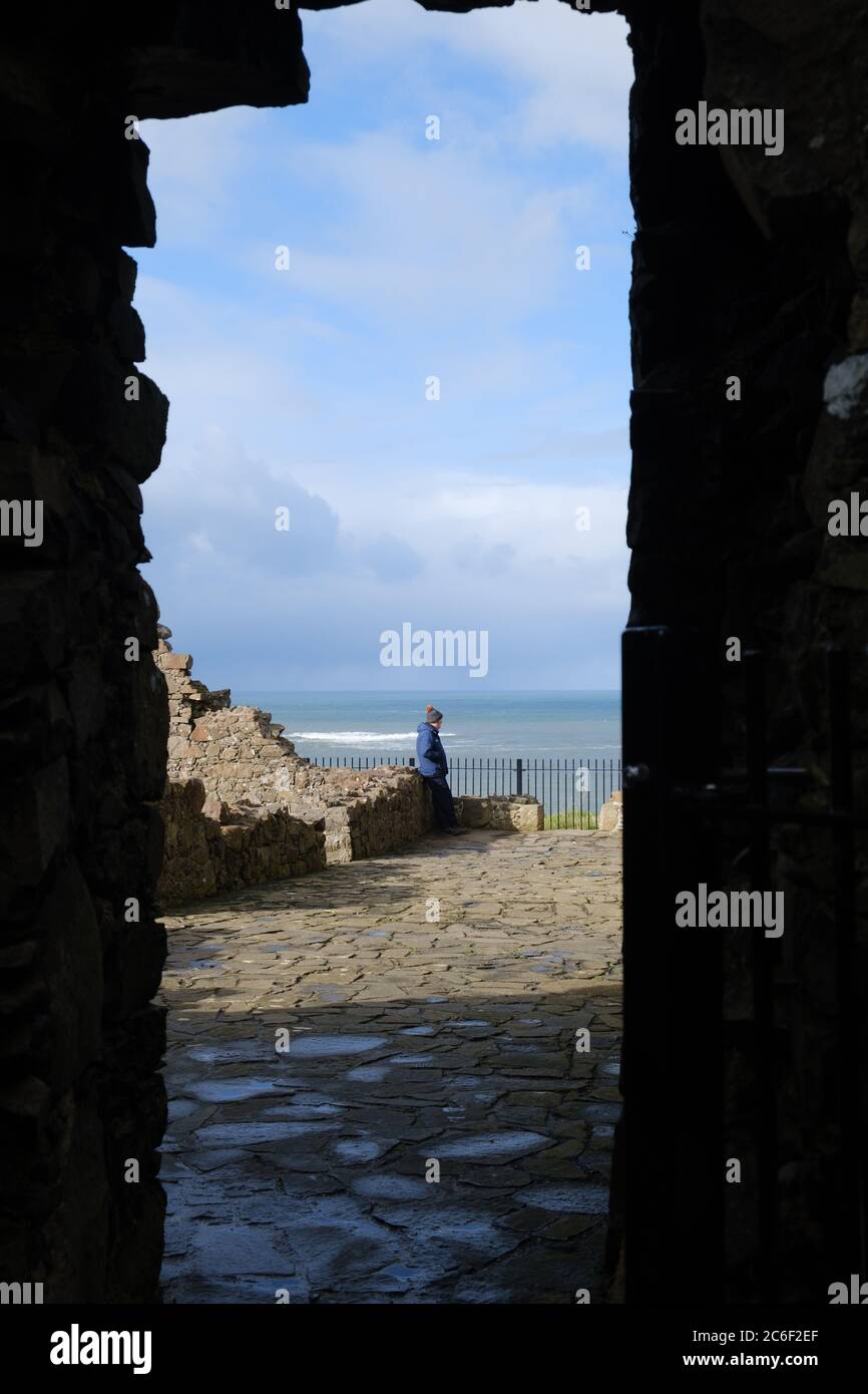 Man in blue coat and bobble hat looks out to sea from the ruins at Dunluce Castle in Co. Antrim, Northern Ireland Stock Photo