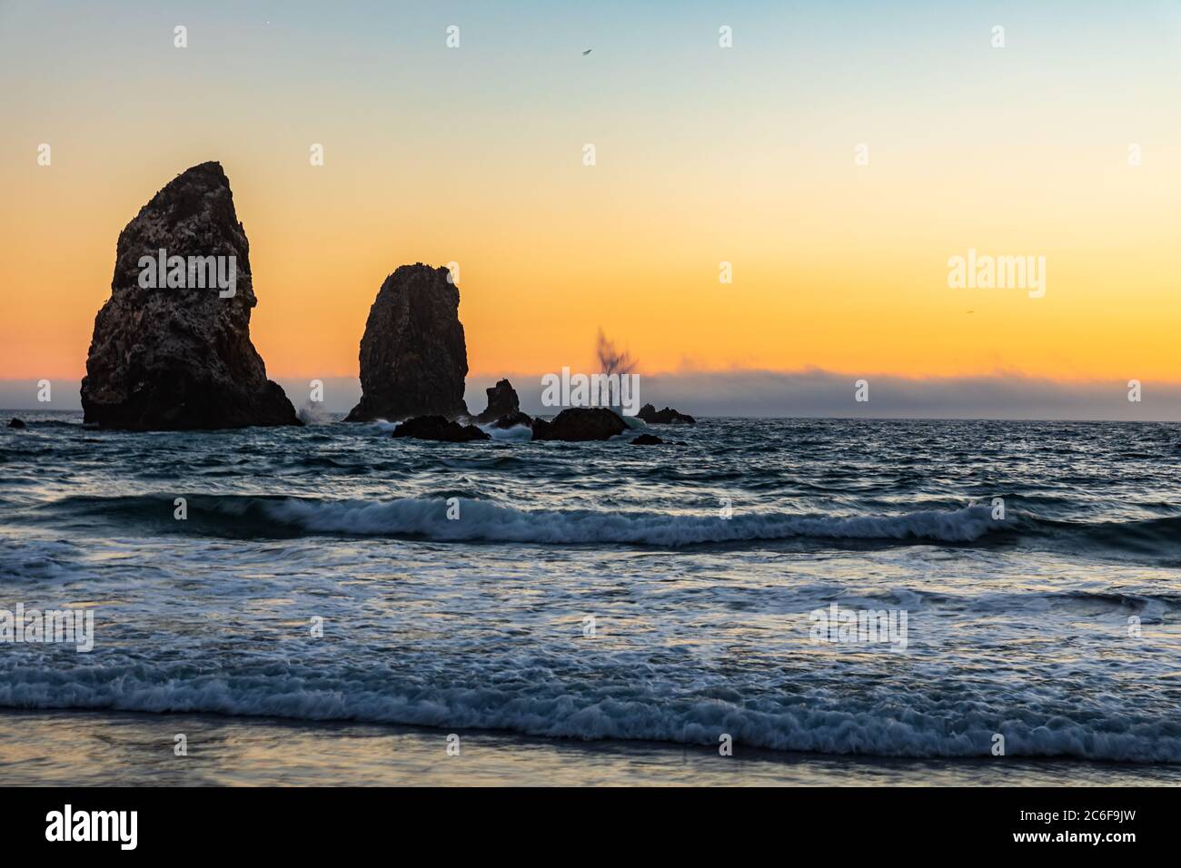 Orange sunset shows silhouette of waves splashing on Sea Stacks on Cannon Beach in Oregon Stock Photo