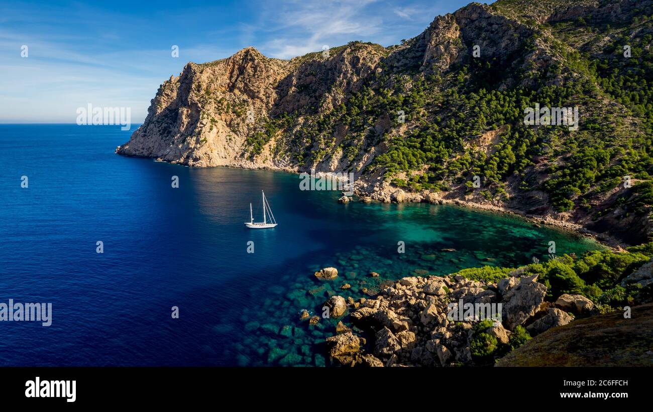 view from torre de cala en basset over a white two-mast sailing boat anchoring in a small idyllic bay at the la trapa mountain ridge nearby sant elm Stock Photo