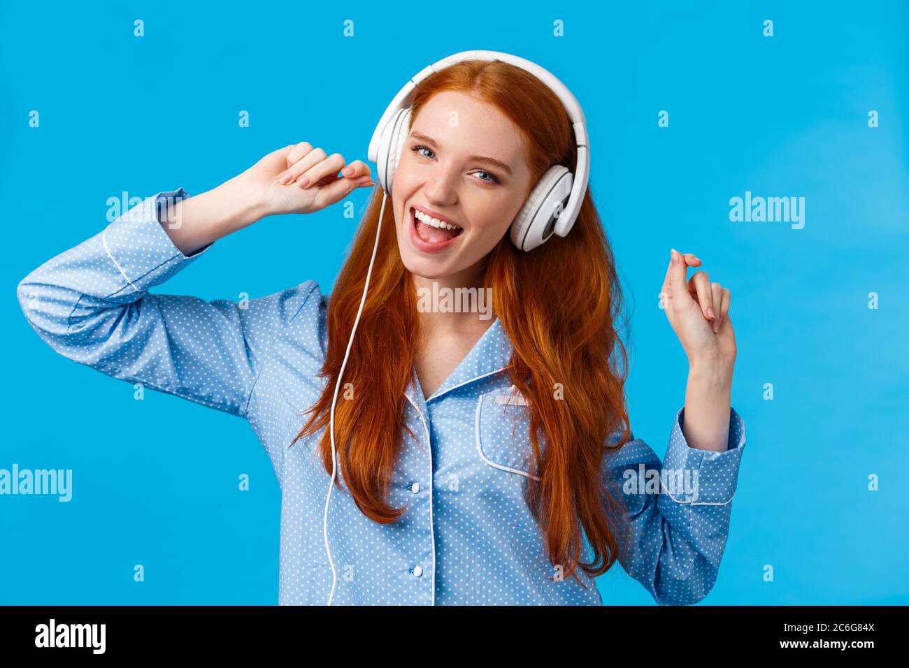 Girl waking up starting day with music. Joyful carefree redhead teenager dancing in nightwear, raising hands up wearing large white headphones singing Stock Photo
