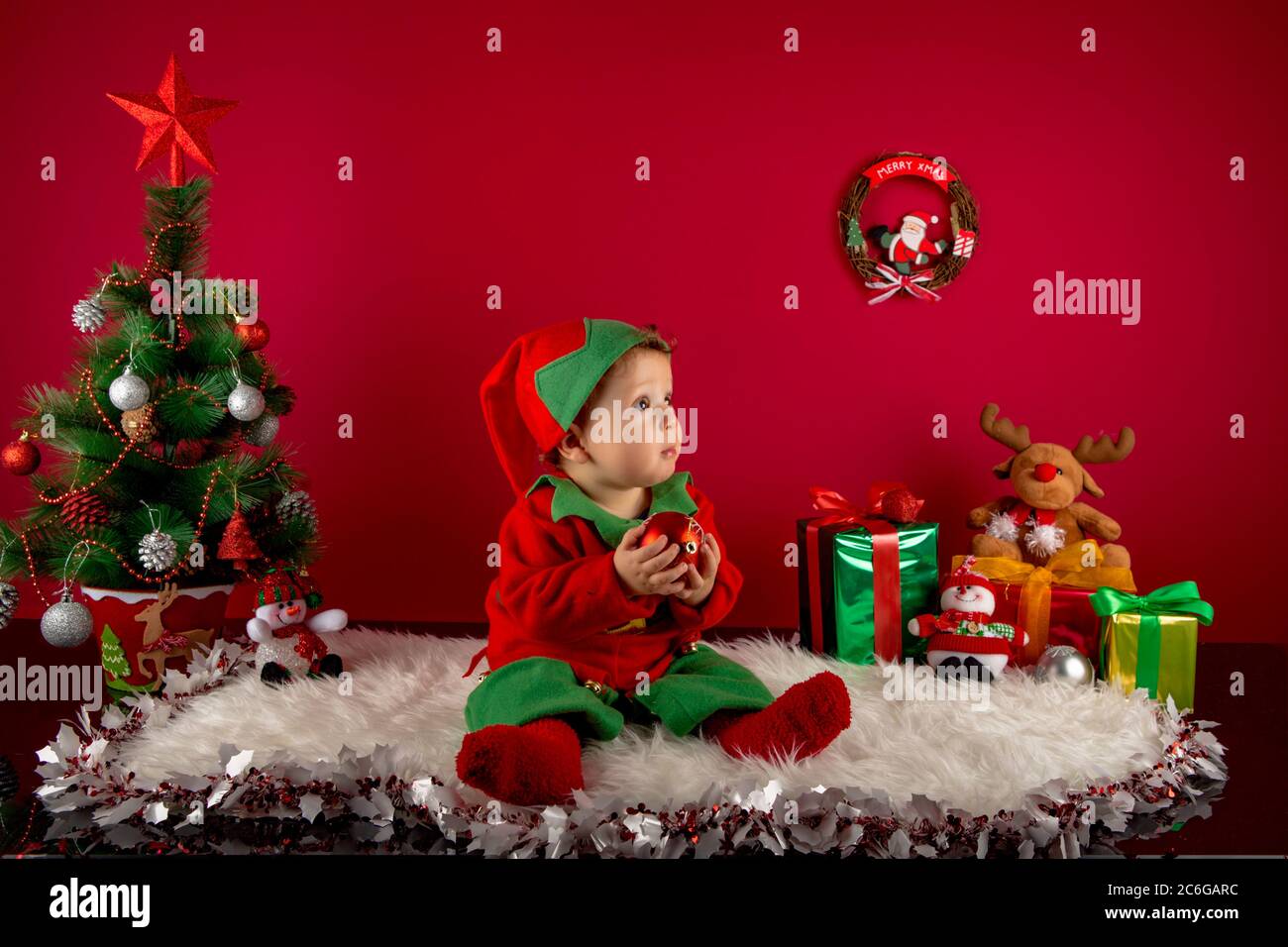 Baby dressed as an elf on her first Christmas, set in a setting with a Christmas tree, gifts, snow, reindeer and decorations Stock Photo