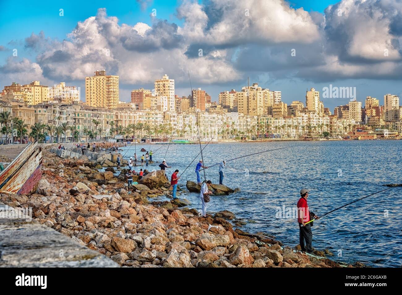 Fishermen on the rocky shoreline of Stanley Bay at sunrise, with the skyline of Alexandria in the background Stock Photo