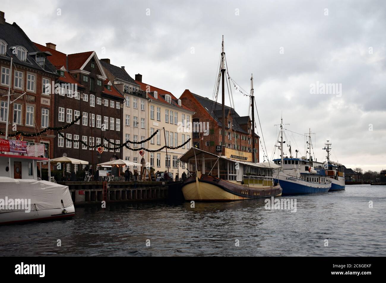 The north side of Nyhavn Canal looking out towards the main canal.  Grey, cloudy skies.  Sail boats are moored against the side of the canal. Stock Photo