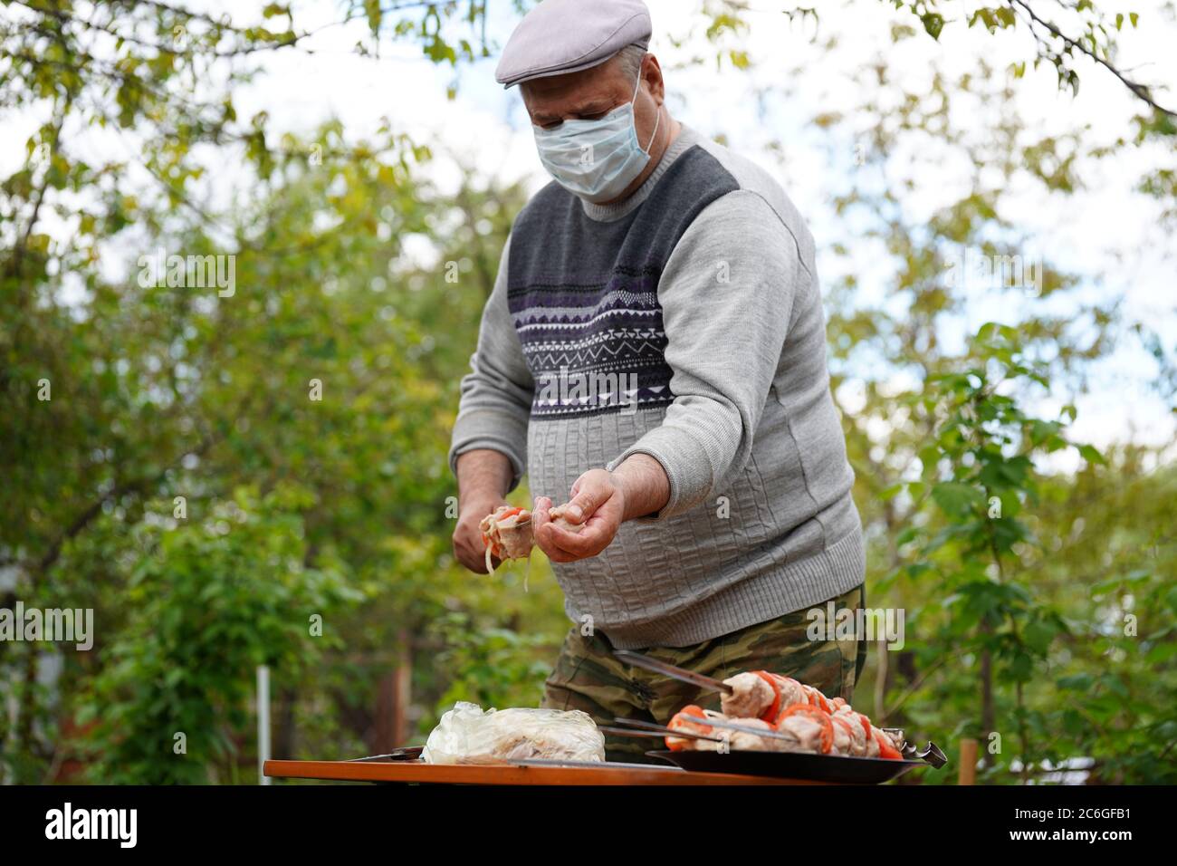 Mature man in medical mask impales raw shashlik on skewer. Older male holds skewer with barbecue. Concept of picnic outdoor. Stock Photo