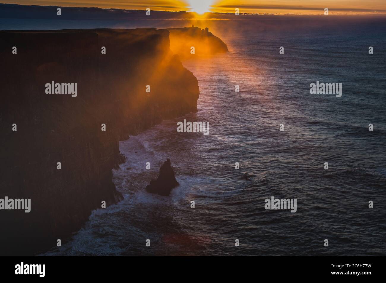 Sun sets on the Cliffs of Moher while mist rises from the Atlantic Ocean. Misty or foggy sunset over dramatic cliffs as sun rays peep through the haze Stock Photo