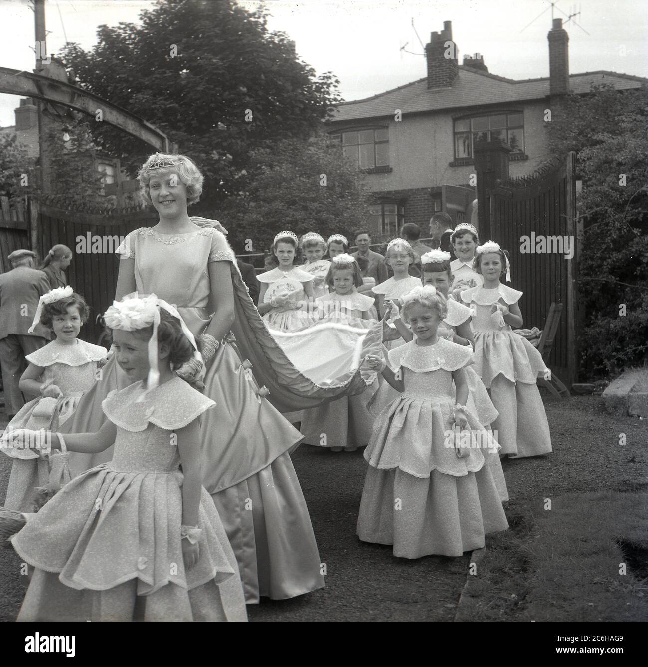 1950s, historical, with a big smile on her face, the town's newly crowned Rose Queen, Farnworth, Bolton, Lancashire, England, UK with her entourage of young girls holding the train, walking in a procession through the local park. Dating back to the 1880s, the annual Rose Queen festival, held in June, became a major annual event in many towns and villages across the UK, especially in Lancashire, known as the red rose county, following the 1455-87 Wars of the Roses. Stock Photo