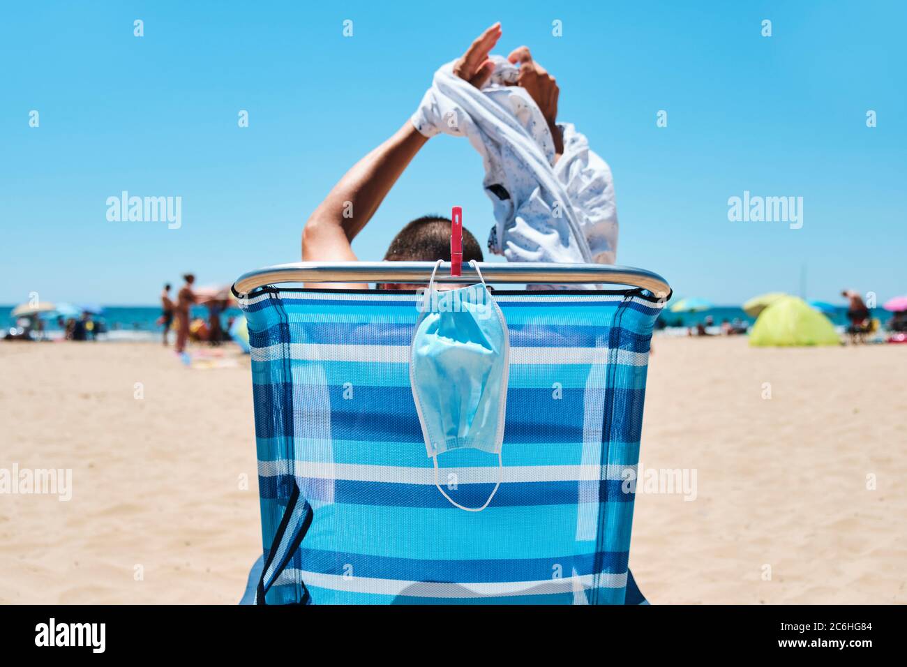 closeup of a caucasian man, sitting in a deck chair on the beach, seen from behind, taking off  his T-shirt while his surgical mask is hanging from th Stock Photo