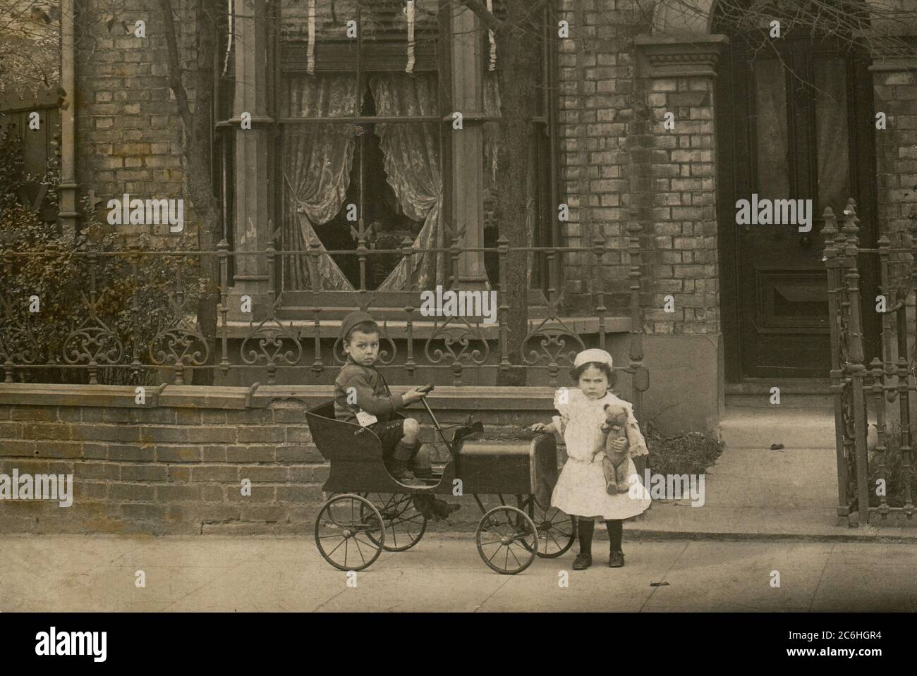 England. Circa 1910. A young boy and his sister are posing for a photograph outside their house. The boy is sitting in a peddle car and has a toy bus conductor's ticket machine slung over his shoulder. The girl is holding a teddy bear. Stock Photo