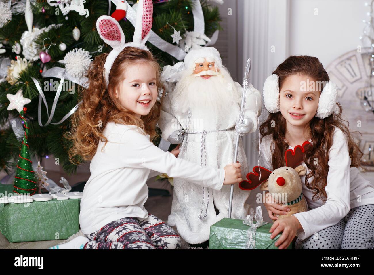 Little girls in comfortable home clothes sitting on floor in beautiful Christmas decorations. Two little sisters decorating Christmas tree with fir-co Stock Photo