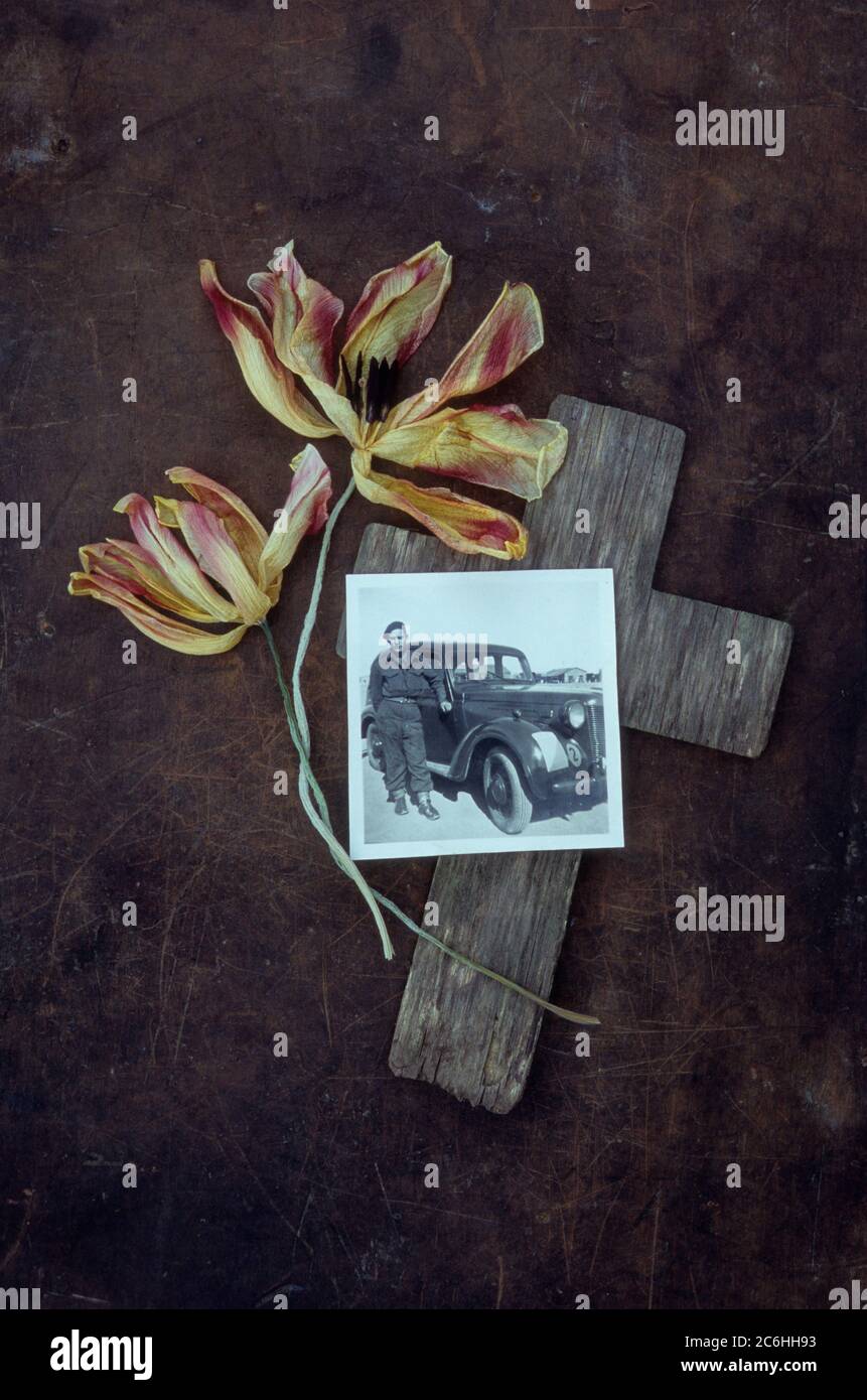 Weathered wooden cross lying on tarnished metal with two dried yellow and red tulips and 1950s photo of army driver and car Stock Photo