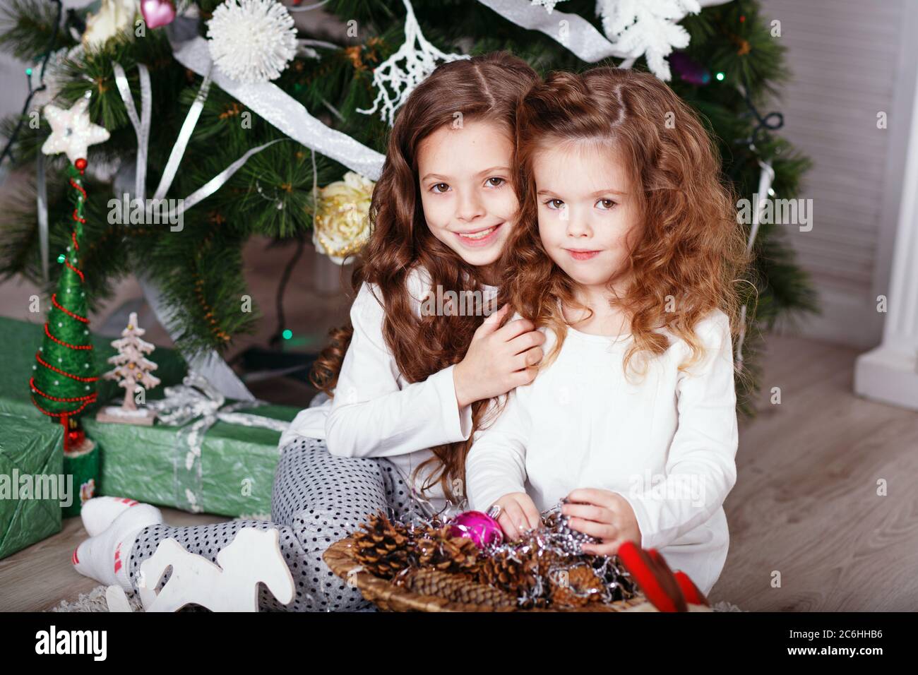 Little girls in comfortable home clothes sitting on floor in beautiful Christmas decorations. Two little sisters decorating Christmas tree with fir-co Stock Photo