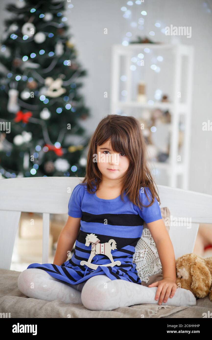 Portrait of pretty brown-eyed little girl in a blue striped dress sitting on a white bench in New Year's interior Stock Photo