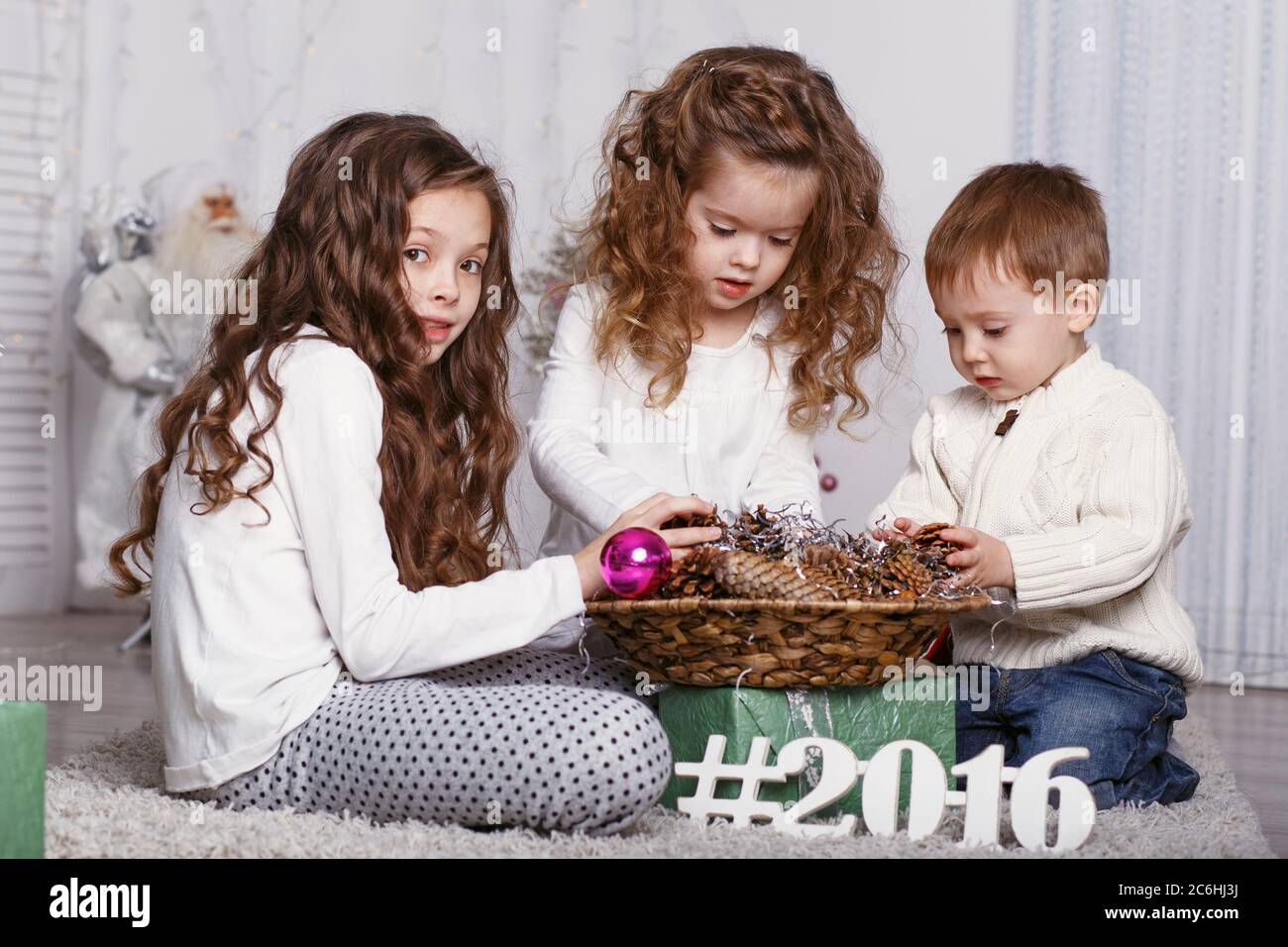 three Little kids in comfortable home clothes sitting on floor in beautiful Christmas decorations. three little siblings decorating Christmas tree wit Stock Photo