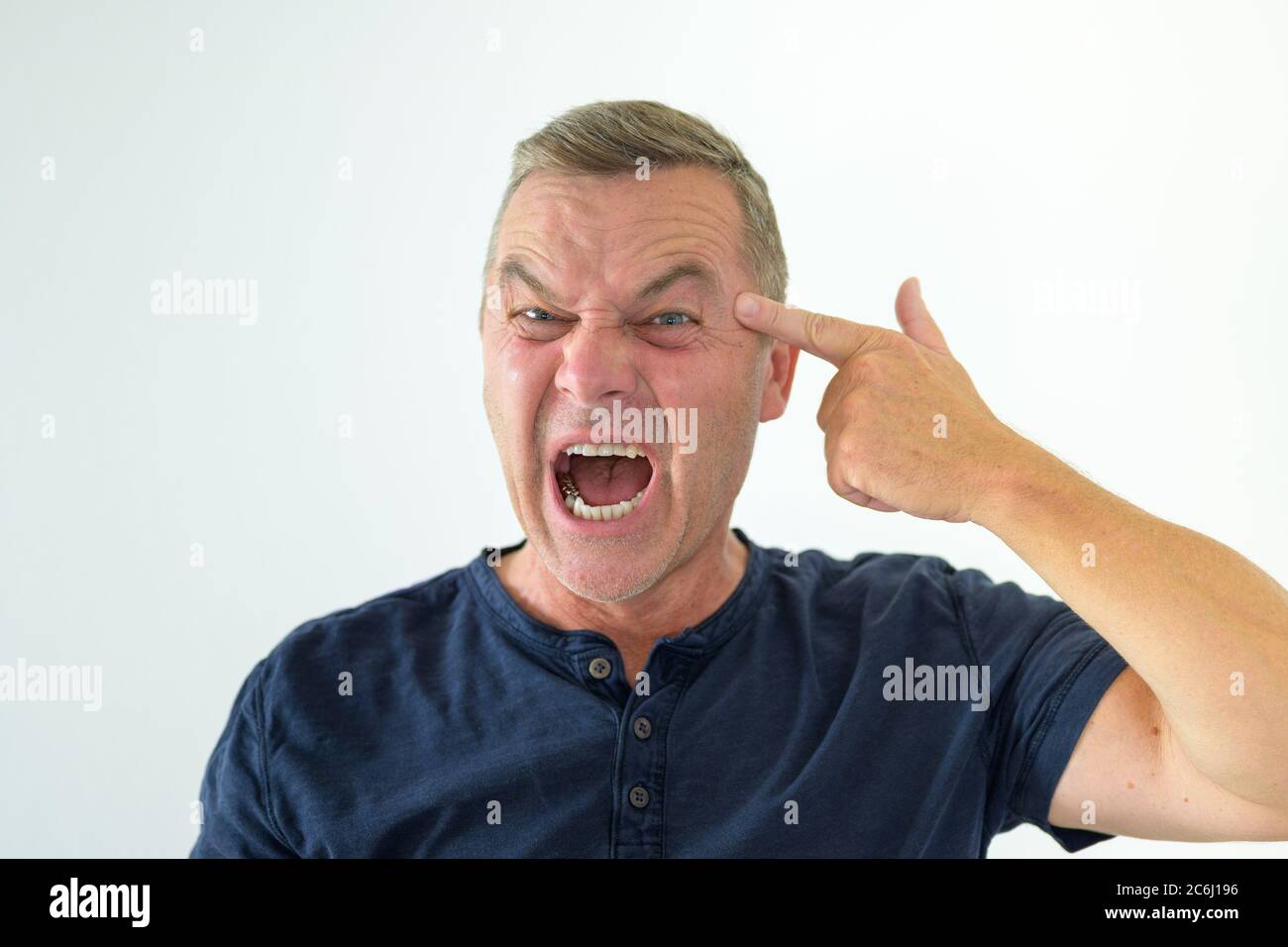 Angry man making a threatening gun gesture with his hand pointing at his own forehead while yelling at the camera in a head and shoulders portrait on Stock Photo