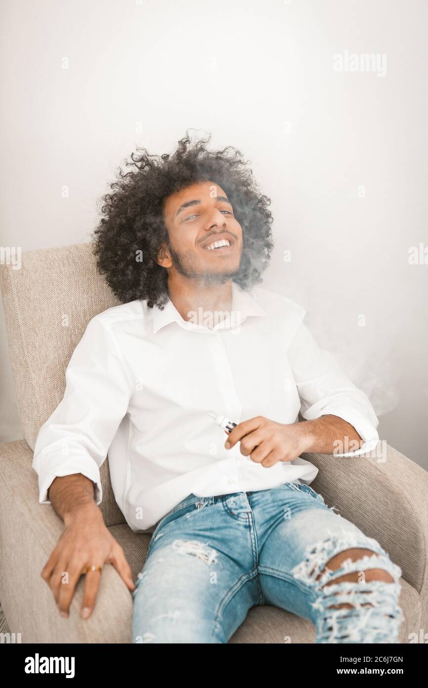 Funny young man sitting and vaping electronic cigarette at home armchair. Young smoker man resting after long working day. Alternative smoking concept Stock Photo