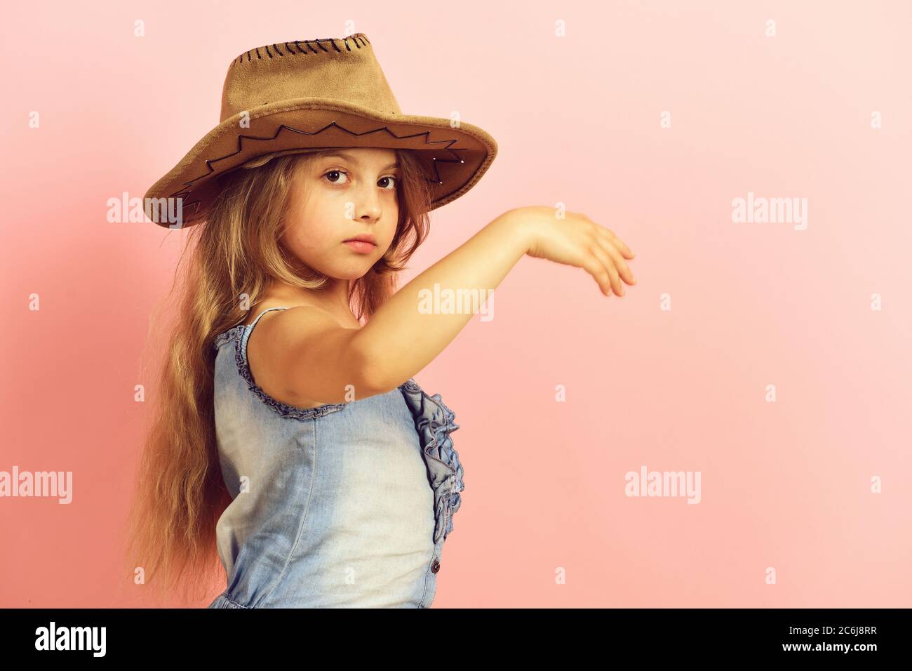 Kid with serious face and long fair hair wears jeans dress. Fashion and  casual style concept. Girl in fancy outfit wears cowboy hat. Little lady in  stylish clothes on pink background, copy