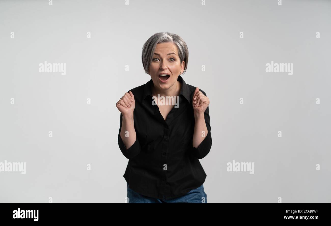 Enthusiastic mature woman with open mouth rejoices in gain. Caucasian lady expresses delight and joy looking at camera while posing in studio on white Stock Photo