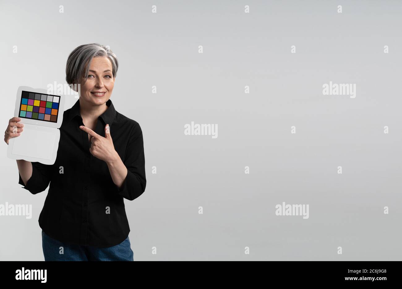Smiling woman holds color checker pointing on it. Gray haired lady in black shirt shows target for camera customization. Mature model isolated on Stock Photo