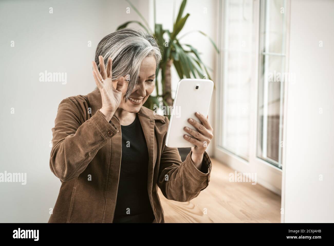 Joyful woman waves her hand using tablet computer for communication with loved ones in internet. Happy aged woman have online conversation on digital Stock Photo