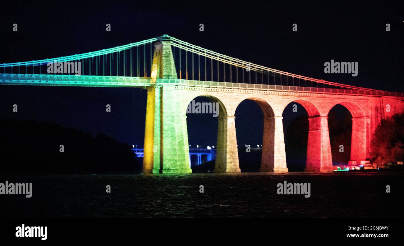 Bangor, Gwynedd, N Wales. Menai suspension Bridge over the Menai Straits is illuminated in rainbow colours to celebrate 72 years of the NHS Stock Photo