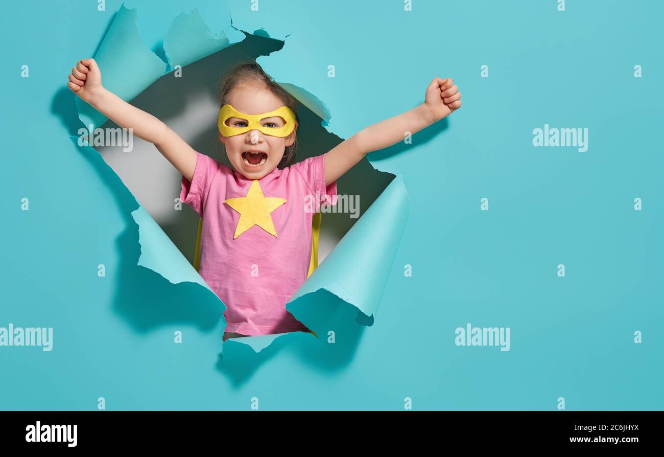 Little child playing superhero. Kid on the background of bright blue wall. Girl power concept. Yellow, pink and  turquoise colors. Stock Photo