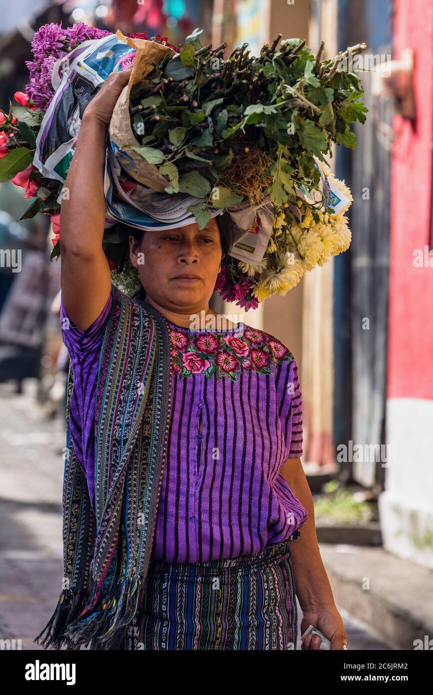 Guatemala, Solola Department, Santiago Atitlan, A Mayan woman wearing traditional dress carries bundles of flowers on her head in the market. Stock Photo