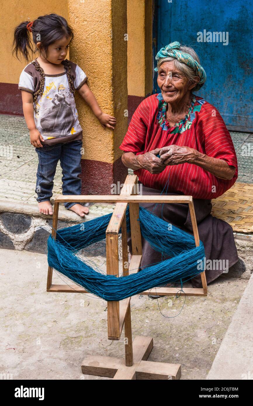 Guatemala, Solola Department, Santa Cruz la Laguna, An older Cakchiquel Mayan woman in traditional dress winds thread on a frame to prepare for weaving on a back loom as a little girl watches. Stock Photo