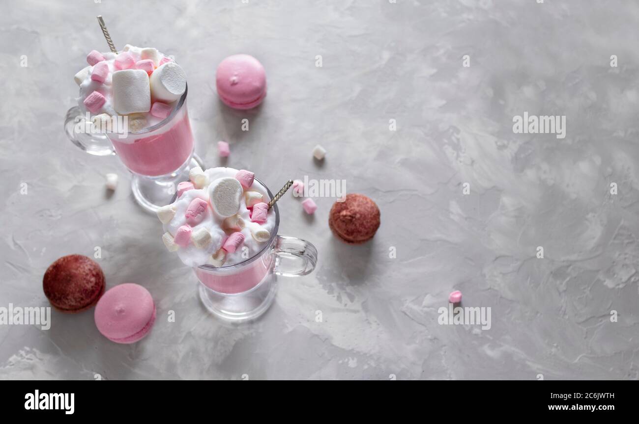 Pink hot chocolate with whipped cream and marshmallow candy in a 2 glass mugs, sweet pastel french macaroons on the grey background. Top view Stock Photo