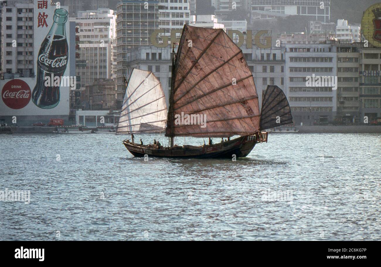 A junk sailing in Victoria Harbour, Hong Kong. 1968. Advertising signs on buildings in the background. Stock Photo