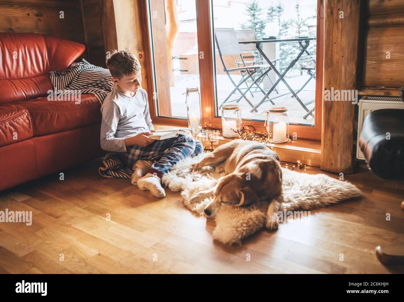 Boy reading book on the floor near slipping his beagle dog on sheepskin in cozy home atmosphere. Peaceful moments of cozy home concept image. Stock Photo