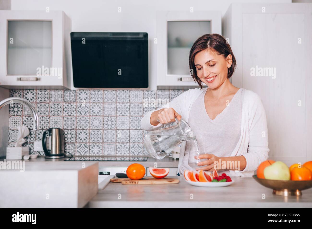 Young sincerely smiling female pouring a purified mineral water into transparent glass decanter at modern kitchen. Plenty of apples, grapefruits, kaki Stock Photo