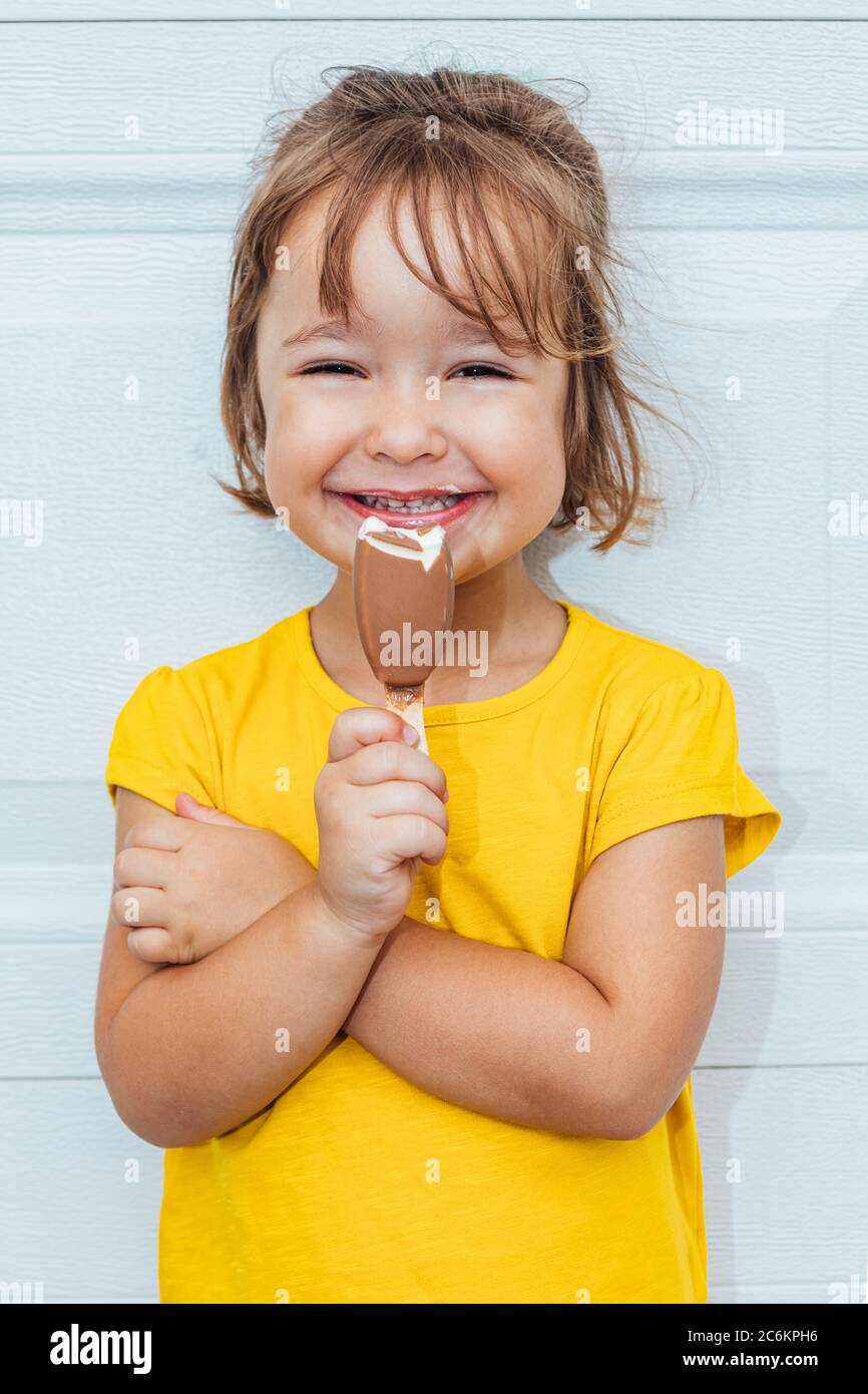 Adorable blond-haired girl eating ice cream, wearing a yellow shirt leaning against white background Stock Photo