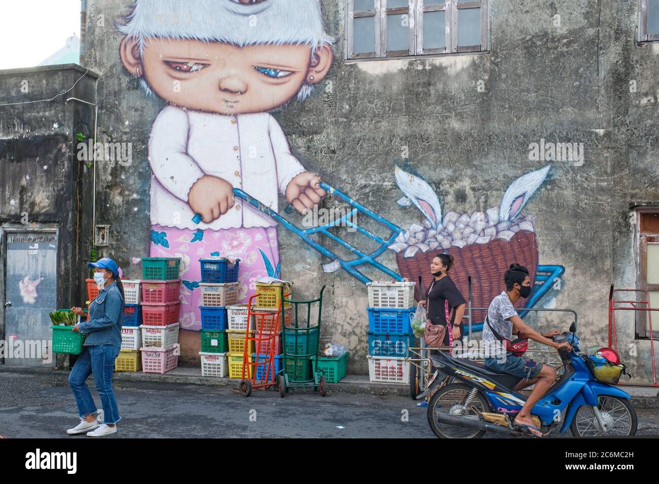 In the market in Phuket Town, Thailand, shoppers pass under a mural with the three-eyed child character named Mardi by Thai street artist Alex Face Stock Photo