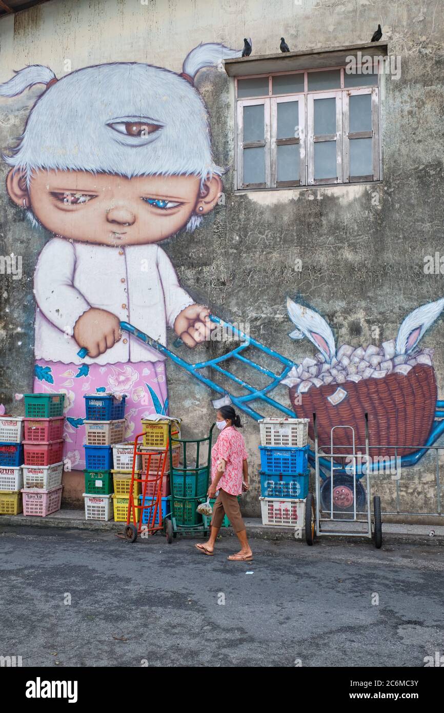 In the market in Phuket Town, Thailand, a woman passes under a mural with the three-eyed child character named Mardi by Thai street artist Alex Face Stock Photo