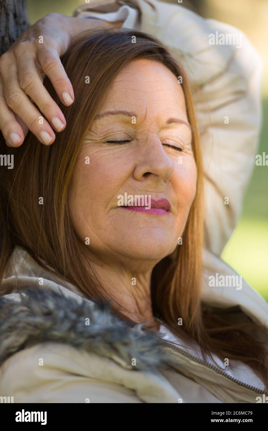 Portrait attractive mature woman relaxed enjoying sunshine in park with closed eyes, daydreaming, contemplating, thoughtful, blurred background. Stock Photo