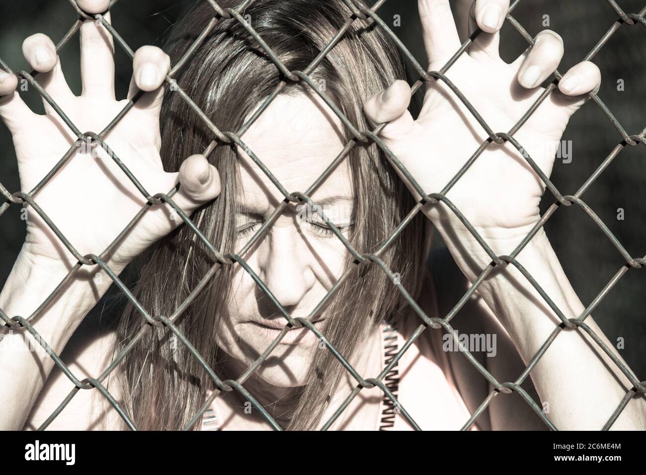 Filtered image Portrait of devastated, stressed mature woman with closed eyes and hands gripped on behind mesh wire fence Stock Photo