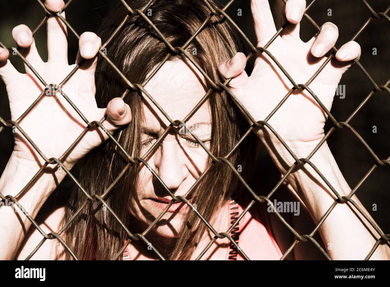 Filtered image Portrait of devastated, stressed mature woman with closed eyes and hands gripped on behind mesh wire fence Stock Photo