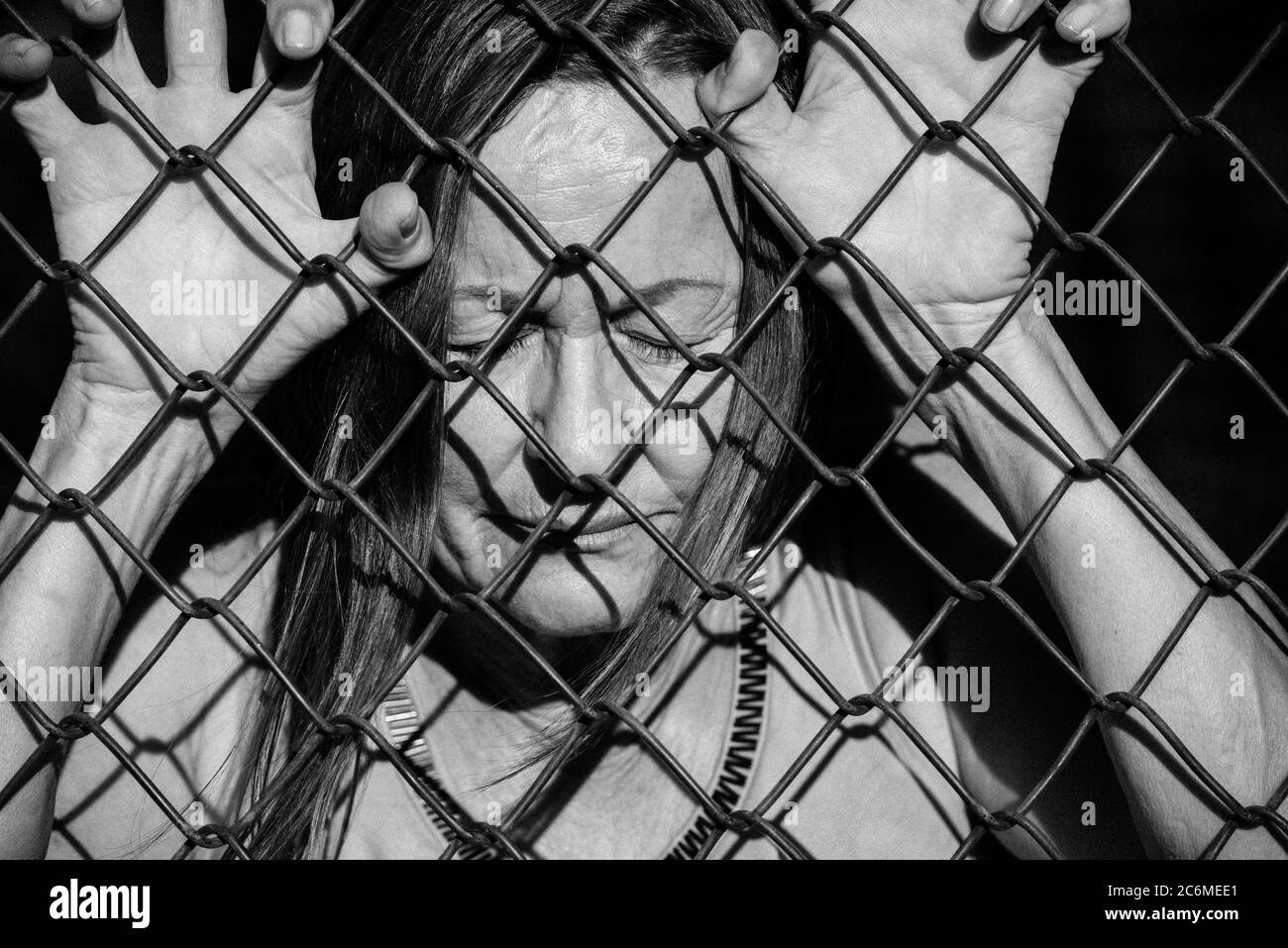 Filtered image Portrait of devastated, stressed mature woman with closed eyes and hands gripped on behind mesh wire fence Stock Photo