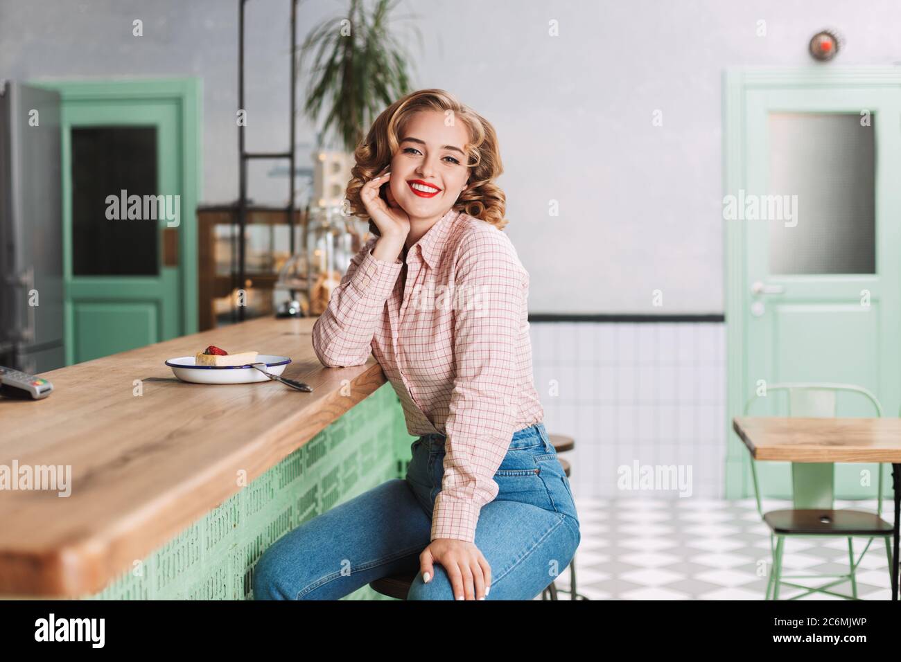 Beautiful lady in shirt and jeans sitting at the bar counter with piece of  cake near and joyfully looking in camera while spending time in cafe Stock  Photo - Alamy