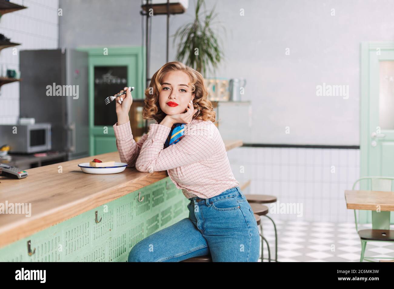 Pretty lady in shirt and jeans sitting at the bar counter in cafe with fork  in hand and cake near and dreamily looking in camera Stock Photo - Alamy