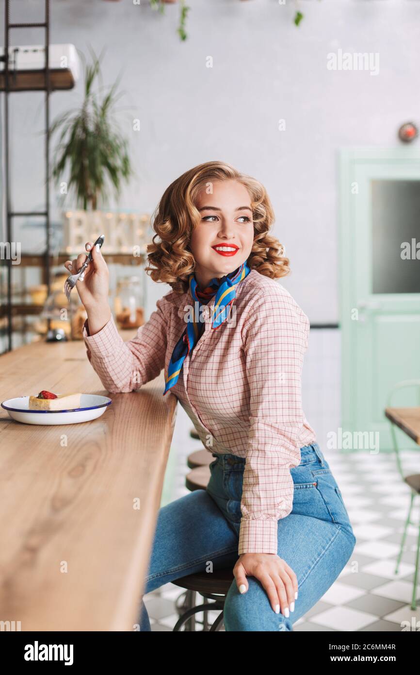 Pretty smiling lady in shirt and jeans sitting at the bar counter in cafe with fork in hand and cake near and dreamily looking aside. Stock Photo