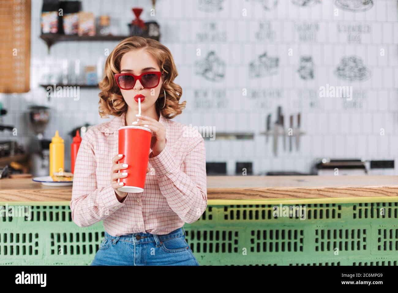 Beautiful lady in sunglasses and shirt sitting at the bar counter and drinking soda water in cafe. Stock Photo