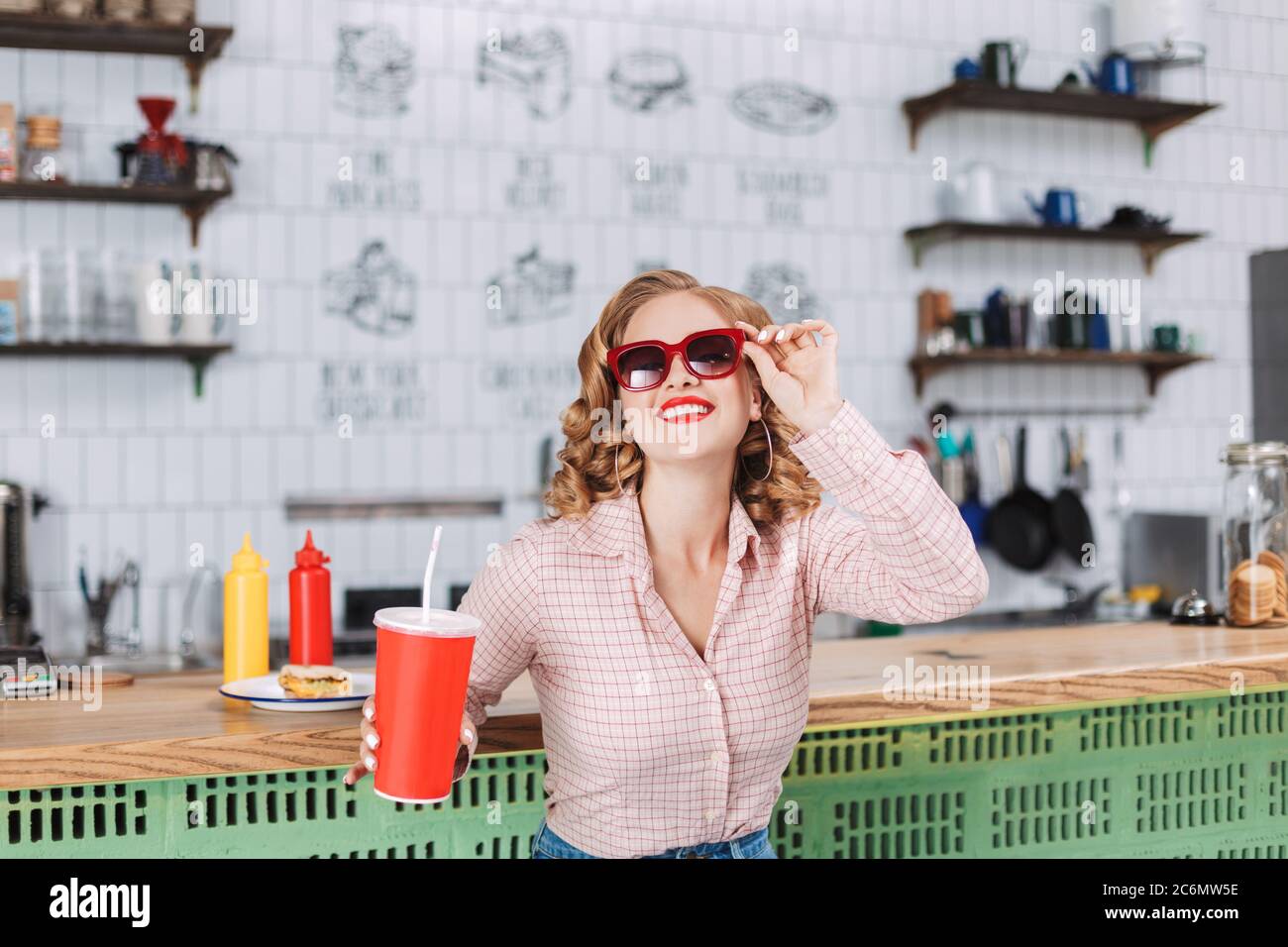 Portrait of smiling lady in sunglasses and shirt sitting at the bar counter with soda water in hand and happily looking in camera in cafe. Stock Photo