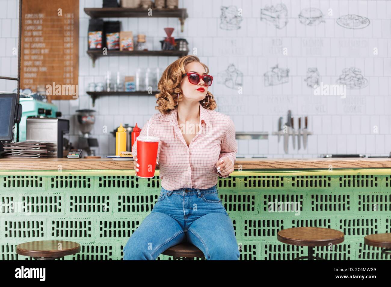 Beautiful lady in sunglasses sitting at the bar counter with soda water in hand and looking aside while spending time in cafe. Stock Photo