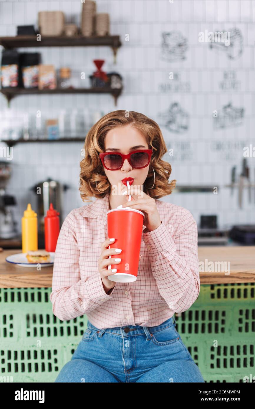 Pretty lady in sunglasses and shirt sitting at the bar counter and drinking soda water while spending time in cafe. Stock Photo
