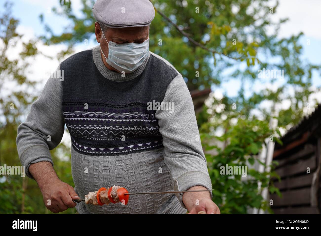 Mature man in medical mask impales raw shashlik on skewer. Older male holds skewer with barbecue. Concept of picnic outdoor. Stock Photo