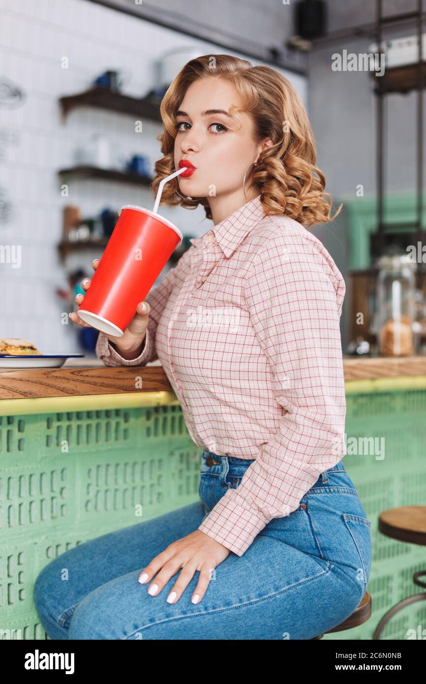 Young beautiful lady in shirt and jeans sitting at the bar counter and drinking soda water while thoughtfully looking in camera in cafe. Stock Photo