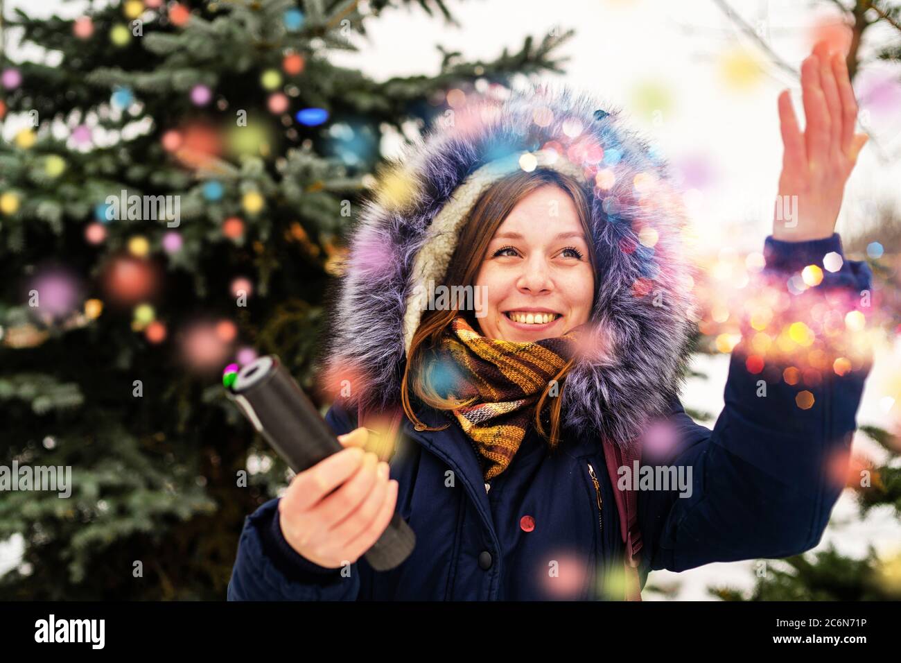 Emotional woman pops confetti petard and smiles with delight. Adult female in hooded jacket admires and reaches out to confetti while standing near Stock Photo